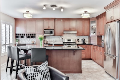 A modern kitchen with wooden cabinetry and stainless steel appliances features a central island with a black countertop. Above the island hang contemporary light fixtures. A vase with tulips sits on the island, adding a touch of color. The kitchen also has a wine rack, a built-in microwave, and a gas stove.