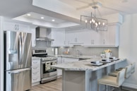 A modern kitchen featuring sleek white cabinetry and stainless steel appliances. The kitchen island has granite countertops with seating for three, set with placemats and glasses. A geometric light fixture hangs over the island, and subtle backsplash tiles add texture to the walls.
