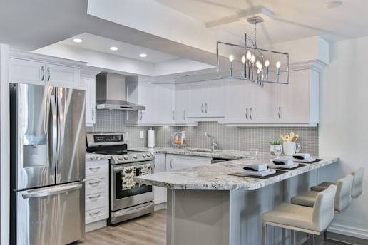 A modern kitchen featuring sleek white cabinetry and stainless steel appliances. The kitchen island has granite countertops with seating for three, set with placemats and glasses. A geometric light fixture hangs over the island, and subtle backsplash tiles add texture to the walls.