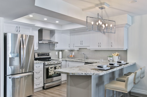 A modern kitchen featuring sleek white cabinetry and stainless steel appliances. The kitchen island has granite countertops with seating for three, set with placemats and glasses. A geometric light fixture hangs over the island, and subtle backsplash tiles add texture to the walls.