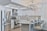 A modern kitchen featuring sleek white cabinetry and stainless steel appliances. The kitchen island has granite countertops with seating for three, set with placemats and glasses. A geometric light fixture hangs over the island, and subtle backsplash tiles add texture to the walls.
