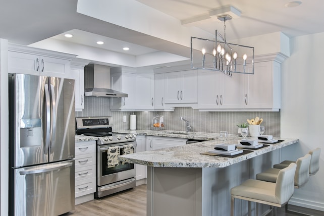 A modern kitchen featuring sleek white cabinetry and stainless steel appliances. The kitchen island has granite countertops with seating for three, set with placemats and glasses. A geometric light fixture hangs over the island, and subtle backsplash tiles add texture to the walls.