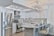 A modern kitchen featuring sleek white cabinetry and stainless steel appliances. The kitchen island has granite countertops with seating for three, set with placemats and glasses. A geometric light fixture hangs over the island, and subtle backsplash tiles add texture to the walls.
