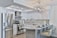 A modern kitchen featuring sleek white cabinetry and stainless steel appliances. The kitchen island has granite countertops with seating for three, set with placemats and glasses. A geometric light fixture hangs over the island, and subtle backsplash tiles add texture to the walls.