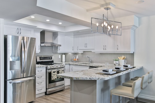 A modern kitchen featuring sleek white cabinetry and stainless steel appliances. The kitchen island has granite countertops with seating for three, set with placemats and glasses. A geometric light fixture hangs over the island, and subtle backsplash tiles add texture to the walls.