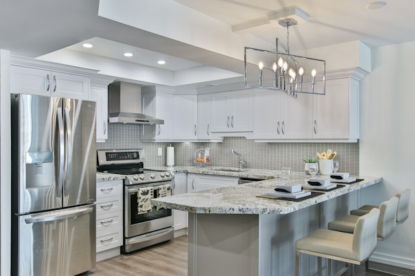 A modern kitchen featuring sleek white cabinetry and stainless steel appliances. The kitchen island has granite countertops with seating for three, set with placemats and glasses. A geometric light fixture hangs over the island, and subtle backsplash tiles add texture to the walls.