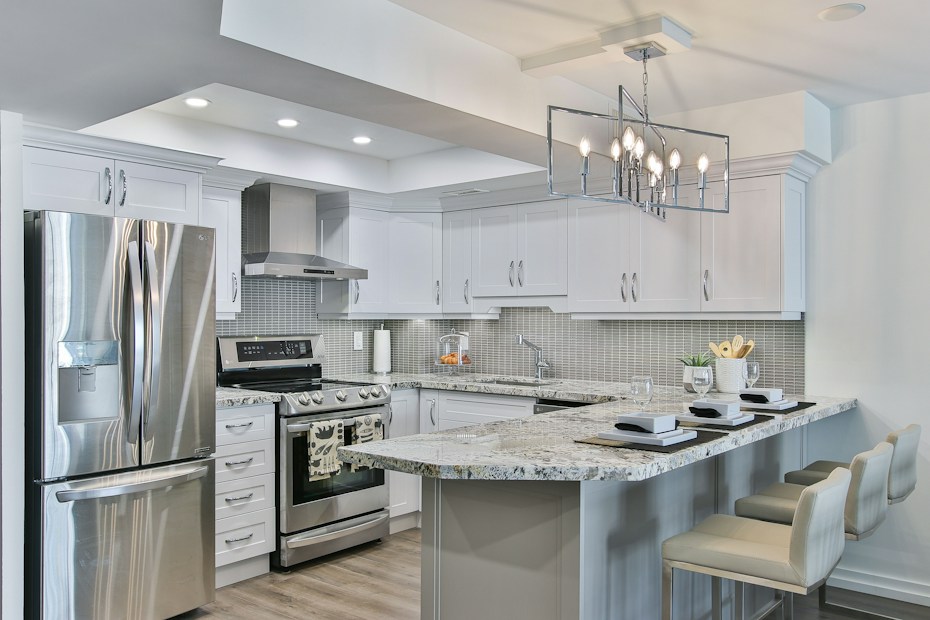 A modern kitchen featuring sleek white cabinetry and stainless steel appliances. The kitchen island has granite countertops with seating for three, set with placemats and glasses. A geometric light fixture hangs over the island, and subtle backsplash tiles add texture to the walls.