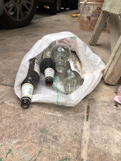 Several empty glass bottles are placed inside a woven white sack on a concrete floor. Nearby, there are car tires, a stool, and a bucket in the background.