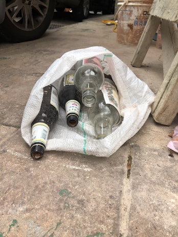 Several empty glass bottles are placed inside a woven white sack on a concrete floor. Nearby, there are car tires, a stool, and a bucket in the background.