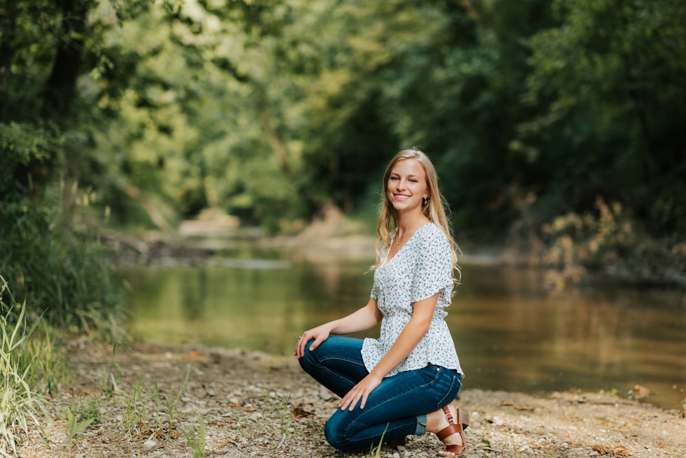 Frau in weiß-schwarzem Blumenhemd und blauer Jeans sitzt auf dem Boden in der Nähe des Flusses