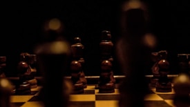 A dimly lit chessboard with wooden chess pieces arranged for a game. The pieces are in shadow, creating a dramatic and mysterious atmosphere. The focus is on the wooden texture and the arrangement suggests an ongoing or paused game.