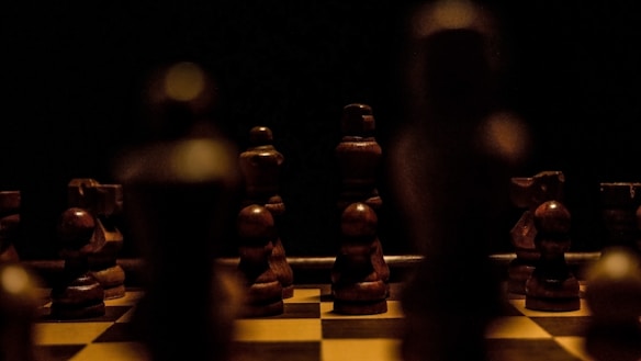 A dimly lit chessboard with wooden chess pieces arranged for a game. The pieces are in shadow, creating a dramatic and mysterious atmosphere. The focus is on the wooden texture and the arrangement suggests an ongoing or paused game.