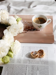 A serene setting featuring a cup of coffee resting on an open magazine. The page displays an image of a woman surrounded by white petals. Several white flowers with green buds partially rest on the magazine, adding a natural touch. On the magazine lies a pair of intertwined, elegant, gold rings, contributing a sense of luxury.