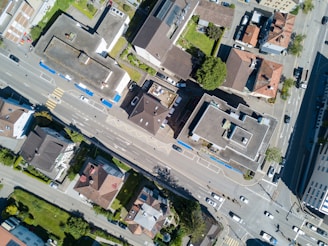 An aerial view of an urban neighborhood with several multi-story buildings, roads with cars, and green areas. The image captures the layout of buildings and streets, showcasing a well-organized city block. The roofs have varied designs, and there are several parking areas visible.