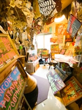 A cluttered room filled with numerous colorful signs and artworks in various languages, leaning against furniture and walls. The ceiling is decorated with hanging papers and fabrics, and a window lets in natural light. The overall scene is chaotic yet vibrant, with an artistic and rebellious atmosphere.