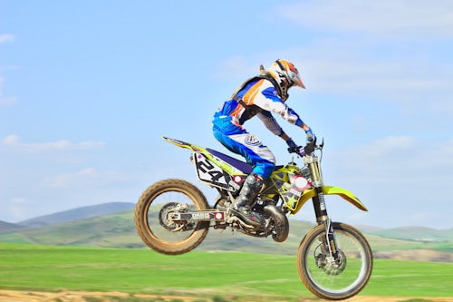 A person wearing a colorful motocross outfit is riding a dirt bike mid-air against a backdrop of green fields and distant hills under a clear blue sky. The bike is in motion, and the wheels are off the ground, indicating a jump or a leap in the air.