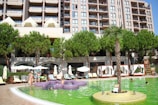 A modern hotel or apartment building with multiple balconies overlooks a lively swimming pool area. The scene includes several people enjoying the pool, lounging on deck chairs under umbrellas, and socializing. There are lush green trees and a palm tree providing shade and decoration around the pool area.