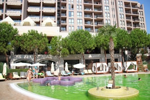 A modern hotel or apartment building with multiple balconies overlooks a lively swimming pool area. The scene includes several people enjoying the pool, lounging on deck chairs under umbrellas, and socializing. There are lush green trees and a palm tree providing shade and decoration around the pool area.