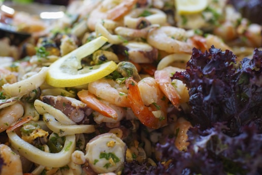 A close-up of a seafood salad featuring a mix of shrimp, mussels, squid, and octopus garnished with lemon slices and herbs. The salad is accompanied by fresh, dark leafy greens, giving it a vibrant and fresh appearance.