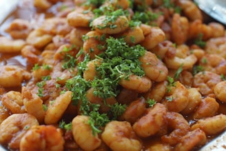 A close-up of a dish featuring cooked shrimp coated in a rich, reddish-brown sauce, garnished with a generous amount of fresh green herbs.