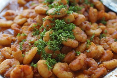A close-up of a dish featuring cooked shrimp coated in a rich, reddish-brown sauce, garnished with a generous amount of fresh green herbs.