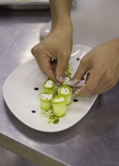 A pair of hands delicately arranging rolled zucchini appetizers filled with creamy cheese on a white oval plate. The dish features small dollops of dark sauce and green herb garnish on a stainless steel surface.