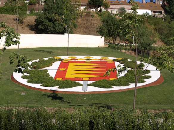 A landscaped garden area features an emblem or crest design made from various plants and colored materials. The design is circular with a red and yellow shield in the center, surrounded by greenery and white gravel. Trees and shrubs encircle the emblem, and a few buildings are visible in the background.
