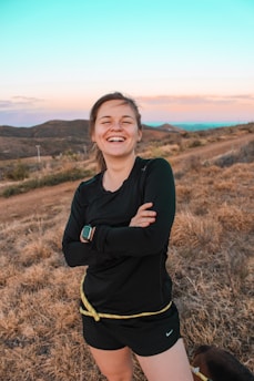 A person is standing outdoors on a grassy hillside, smiling broadly with arms crossed. They are wearing a black athletic outfit with a smartwatch on their wrist. The background features rolling hills and a distant horizon under a sky transitioning from sunset hues to blue.