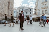 A bustling urban street scene with people walking and interacting in front of historic architecture. The scene includes an older man in traditional attire holding a cane, a cyclist wearing a blue jacket, and other pedestrians. The background features detailed stone buildings and outdoor cafes, contributing to a lively city atmosphere.
