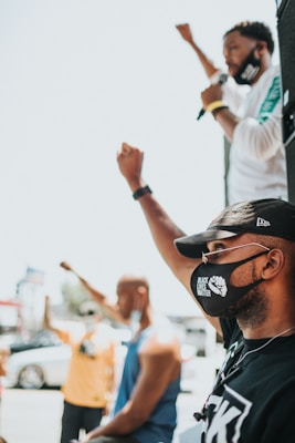 A group of individuals are raising their fists in a gesture of solidarity. One person, wearing a face mask with the text 'Black Lives Matter,' is prominently in the foreground, while another individual is speaking into a microphone. The scene appears to be outdoors, and the image captures a moment of protest or activism.