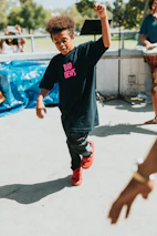 A child is energetically dancing outdoors, wearing a black T-shirt with the words 'BAD NEWS' in pink and red sneakers. A person plays a drum in the background, and a microphone stand is visible. The setting appears to be a park area with greenery.