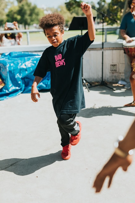 A child is energetically dancing outdoors, wearing a black T-shirt with the words 'BAD NEWS' in pink and red sneakers. A person plays a drum in the background, and a microphone stand is visible. The setting appears to be a park area with greenery.