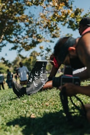 A person sitting on grass, holding a sign with 'WE THE MOVEMENT' written on it. Another person nearby with a camera, possibly taking a picture. In the background, people are gathered under a tree with green leaves, with a clear blue sky above.