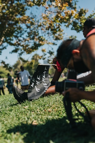 A person sitting on grass, holding a sign with 'WE THE MOVEMENT' written on it. Another person nearby with a camera, possibly taking a picture. In the background, people are gathered under a tree with green leaves, with a clear blue sky above.