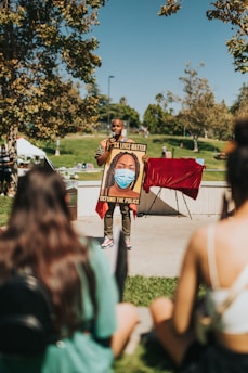 A person is standing outdoors holding a poster during a gathering. The poster includes the text 'Black Lives Matter' and 'Defund the Police,' along with an illustration of a person wearing a mask. Trees and grass are visible in the background, along with blurred figures sitting in the foreground.