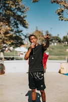 A person holding a microphone is standing outdoors on a sunny day. They are gesturing with one hand as if speaking or performing. Trees and a park with sunlit grass are visible in the background, along with a few tents.