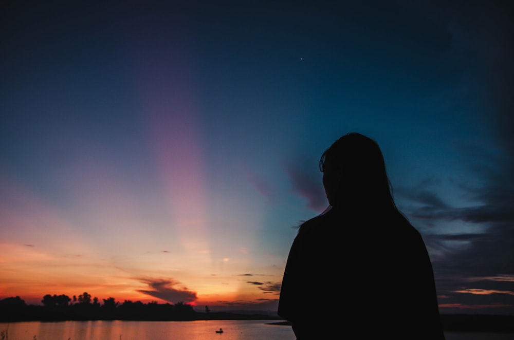 silhouette of man standing near body of water during sunset