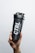 A hand is holding a black shaker bottle with the text 'CTRL A MEAL REPLACEMENT' printed on its side. The background is plain and light gray, providing a contrast to the dark color of the bottle.