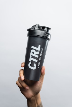 A hand is holding a black shaker bottle with the text 'CTRL A MEAL REPLACEMENT' printed on its side. The background is plain and light gray, providing a contrast to the dark color of the bottle.