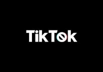 A minimalist logo featuring the word TikTok in a bold, white font centered on a black background. The 'o' in TikTok is stylized with a design resembling a musical note, incorporating subtle blue and red hues.