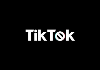 A minimalist logo featuring the word TikTok in a bold, white font centered on a black background. The 'o' in TikTok is stylized with a design resembling a musical note, incorporating subtle blue and red hues.
