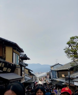 A bustling street filled with people, bordered by traditional Japanese wooden buildings with tiled roofs on either side. In the background, distant mountains and a cloudy sky can be seen. The image captures the essence of a busy cultural or historical area, possibly a market or tourist destination.