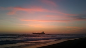 A serene seascape with a large cargo ship silhouetted against a vibrant sunset. The sky is painted with hues of orange, pink, and blue, creating a peaceful and calming atmosphere. Gentle waves are visible on the beach, enhancing the tranquil feeling.