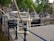 A canal lock mechanism with metal and wooden components is situated by a cobblestone path, surrounded by railings. There are trees and traditional European architecture, including a row of brick buildings with large windows and outdoor seating areas. A pigeon is seen resting near the canal edge.