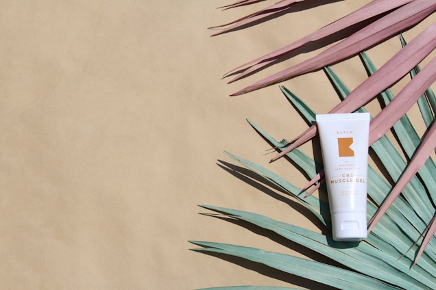 A tube of CBD muscle gel is placed on a flat surface, surrounded by teal and pink palm leaves arranged artistically. The background is a light beige color, providing contrast to the green and pink shades of the palm leaves.