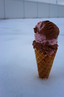 An ice cream cone with two scoops, chocolate and pink in color, stands upright in the snow. The background consists of a blurred fence and sky, creating a stark contrast with the snow and ice cream.