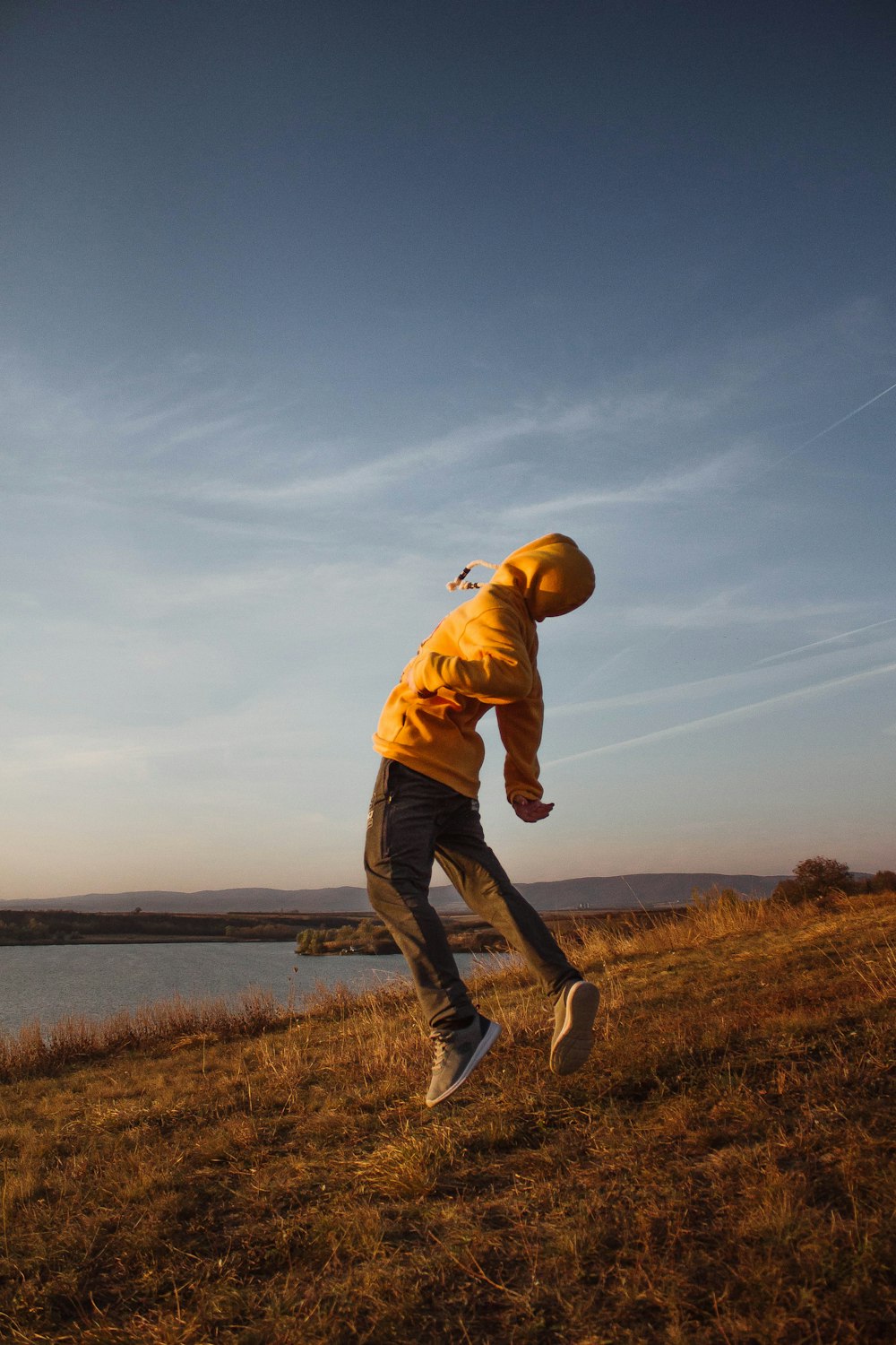 man in brown hoodie and blue denim jeans standing on grass field near body of water