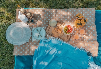 A cozy outdoor picnic setup on a polka-dot mat placed over grass. The arrangement includes a woven hat, two plates with cutlery, a wooden platter with crackers, cheese, and a bowl of strawberries and grapes. Two glasses of juice and a floral arrangement of yellow flowers add to the inviting atmosphere.