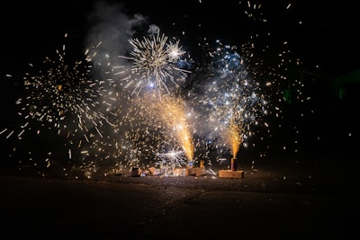 Bright and colorful fireworks erupt against a dark night sky, creating vibrant streaks and bursts of light. The scene includes multiple fireworks set on the ground, with visible sparks and smoke patterns.