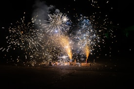 Bright and colorful fireworks erupt against a dark night sky, creating vibrant streaks and bursts of light. The scene includes multiple fireworks set on the ground, with visible sparks and smoke patterns.